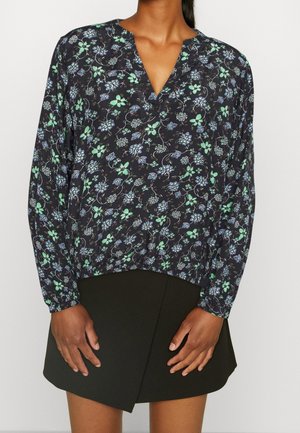 Woman wearing a black floral long-sleeve blouse with green and blue flowers and a black wrap-style mini skirt.