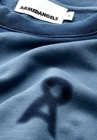 Blauwe kleding met een "ARMEDANGELS" label en maatlabel, met een subtiele donkere abstracte logoafdruk op de stof nabij de kraag.