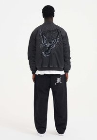Black bomber jacket with embroidered eagle and '97' design on the back; paired with loose black jeans featuring a white logo on the pocket.