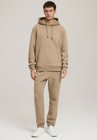 Beżowy zestaw bluzy i spodni dresowych z miękkiego materiału, z przednią kieszenią, sznurkami i ściągaczami, w połączeniu z białymi sneakersami.