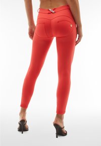 Leggings rossi con una vestibilità aderente, caratterizzati da tasche finte e una superficie liscia. Abbinati a sandali con tacco alto per creare un contrasto.