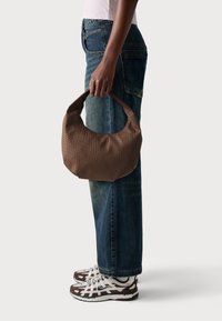 Brun vävd handväska med rundad form och stadigt handtag, hålls vid sidan, kombinerad med högmidjade blåjeans och mönstrade sneakers.