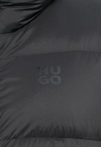 Čierna prešívaná páperová bunda s matným povrchom, so štruktúrovanou nášivkou s logom "HUGO" v vyrazenom čiernom písme.