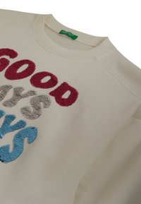 Sudadera crema con texto de lentejuelas multicolores que dice "SOLO BUENAS VIBRACIONES" en letras rojas, beige y azules, con una etiqueta de marca verde en el interior del cuello.