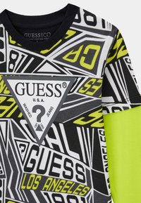 Grafisches T-Shirt mit auffälligen schwarzen, weißen und neongelben Mustern, einem schwarzen, gerippten Rundhalsausschnitt und einem dreieckigen Logo-Patch auf der Vorderseite.