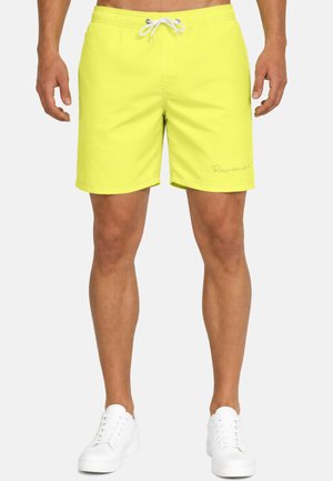 Uomo con pantaloncini da bagno giallo acceso con coulisse bianca e sneakers bianche, in piedi davanti a uno sfondo semplice.