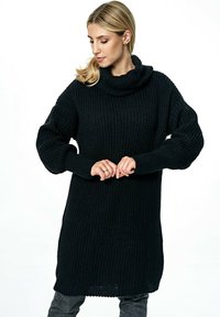 Czarny dzianinowy sweter z wysokim, złożonym kołnierzem, luźnym, teksturowanym korpusem i długimi rękawami. Dół swetra jest nieco dłuższy z tyłu.