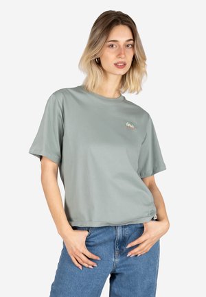 Young woman with shoulder-length blonde hair wearing a light green oversized T-shirt with a small rainbow design and blue jeans.