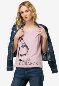Camiseta de cuello en V rosa con arte gráfico de un pingüino y el texto "LAURASØN". Combinada con una chaqueta de mezclilla azul, mostrando un estilo informal.