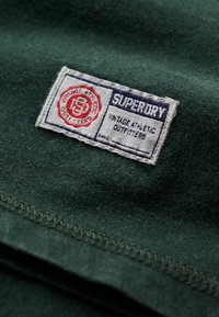 Grøn stof med et mærke med "Superdry" og "Vintage Athletic Outfitters," røde accenter og hvid søm i kanterne.
