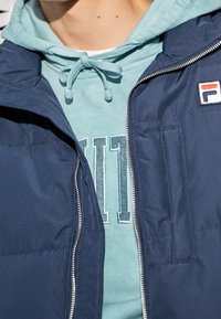 Marinblå zip-jacka med en texturerad yta, bärs över en ljusgrön hoodie med en tryckt logotyp. Synlig dragsko på hoodien.