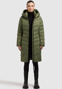 Olivengrøn dunjakke med hætte, quiltet design, lynlås foran, to sidelommer og mærke; båret over et sort outfit og støvler.