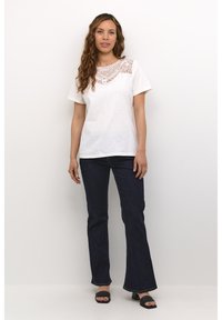 White short-sleeve t-shirt featuring intricate lace detailing at the neckline, paired with dark denim flared pants and black open-toe footwear.