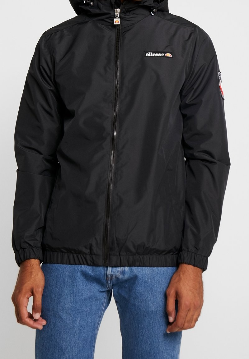 Homme portant une veste noire à capuche zippée avec le logo ellesse sur la poitrine et un patch sur le bras, associée à un jean bleu.