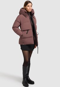 Chaqueta acolchada en malva con un cuello alto y cordón ajustable. Presenta un cierre de cremallera frontal, bolsillos laterales y una silueta cuadrada. Combinada con un vestido corto negro.