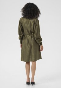 Femme aux cheveux bouclés portant une robe texturée olive à mi-longueur et des chaussures à talons hauts noires, se tenant de dos sur un fond uni.