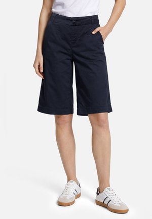 Persona che indossa pantaloncini blu scuro fino al ginocchio, scarpe da ginnastica bianche con strisce nere e una maglietta bianca, in piedi con una mano in tasca.