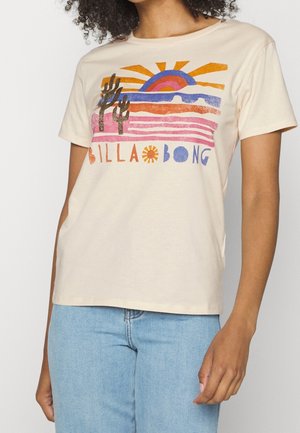 Kobieta w beżowym t-shircie z kolorowym zachodem słońca na pustyni, grafiką kaktusów i napisem „Billabong”, zestawiona z jasnoniebieskimi dżinsami.