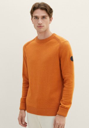 Orange knit sweater with ribbed neckline and cuffs. Features a circular logo patch on the left sleeve. Smooth texture, regular fit.