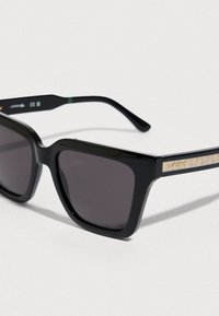 Μαύρα ορθογώνια γυαλιά ηλίου με γκρι φακούς, διακοσμημένα με υφή χρυσού στοιχείου στον βραχίονα με το λογότυπο της μάρκας. Πλαστικός σκελετός και κομψός σχεδιασμός.