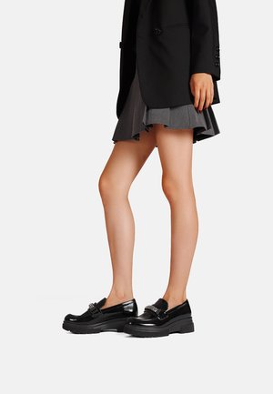 Mocasines de charol negro con una suela gruesa y texturizada, y detalle decorativo de hardware. Combinados con una falda gris plisada y un blazer negro.