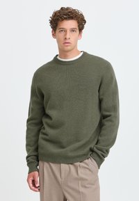 Junger Mann mit lockigem Haar trägt einen olivgrünen Pullover und beige Hose, steht vor einem schlichten hellen Hintergrund.