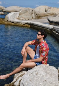 Man met een zonnebril, een rood bloemenhemd en blauwe shorts zit blootsvoets op rotsen bij helder water onder een zonnige hemel.
