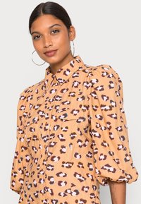 Camisa em pêssego com um padrão de estampado de leopardo; apresenta frente com botões, dois bolsos no peito e mangas bufantes para uma textura adicional.