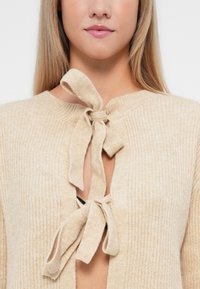 Bežs adīts kardigans ar apaļu dekoltē, atvērtu priekšpusi ar sasietu loku un strukturētu rakstu visā audumā.