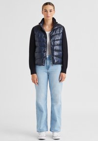 Marineblauer Steppwesten mit gewebtem Design und hohem Kragen, kombiniert mit hellblauen weiten Jeans und weißen Turnschuhen. Glatte Textur, Frontreißverschluss.