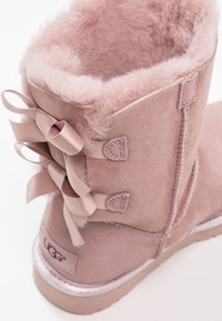 Bottine en suédine rose avec intérieur moelleux, ornée de bandes en satin à l'arrière et d'une semelle en caoutchouc. Détails de couture simples visibles.