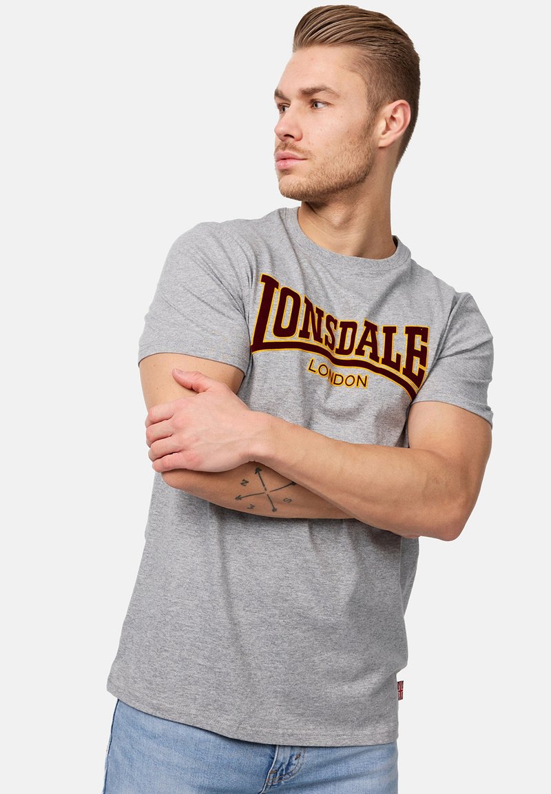 Pilkas medvilnės marškinėlis su ryškiu bordo "LONSDALE" logotipu, trumpomis rankovėmis ir apvalia iškirpte, derinamas su šviesiai mėlynomis džinsomis.