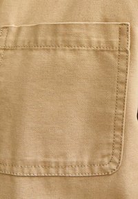 Beige stof met een opvallende klepzak. Getextureerd canvasmateriaal en zichtbare stikaccenten benadrukken de duurzame constructie.