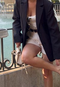 Femme portant un blazer sombre et une tenue blanche, tenant un sac beige, ajustant sa chaussure près d'une rambarde métallique ornée près d'une fontaine.