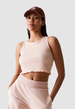 Geribbelde, lichtroze crop top met een scallop zoom en witte logo detail; gecombineerd met bijpassende lichtroze joggers. Bruine pet accentueert de look.