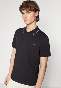 Black polo shirt in cotton blend, featuring a classic collar, three buttons, and white trim on the collar and sleeves. Brand logo on chest.