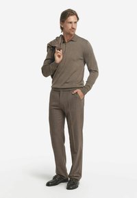 Braunes Polohemd, kombiniert mit einem passenden Pullover, der über die Schulter gelegt ist, ergänzt durch hellbraune Hosen und schwarze Schnürschuhe.