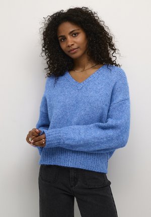 Pull en tricot bleu clair avec un décolleté en V, des manches larges, des poignets et un ourlet côtelés. Associé à un jean gris anthracite. Texture douce et confortable.