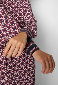 Mains ajustant le poignet d'une chemise à manches longues à motif géométrique rouge, blanc et bleu avec des poignets rayés.