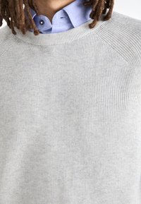 Ljusgrå räfflad tröja med rund halsringning, som bärs över en ljusblå skjorta med krage. Tyget verkar mjukt med en subtil textur.