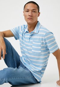 Polo w paski w jasnoniebieskim i białym kolorze, wykonany z dzianiny, krótki rękaw, kołnierzyk i jedna kieszeń na piersi. Noszony z niebieskimi dżinsami.