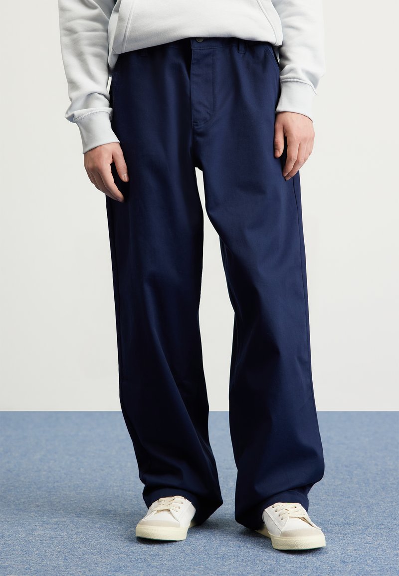 Pantaloni a gamba larga blu navy con una texture liscia, chiusura frontale con bottone e tasche laterali, abbinati a sneaker color crema.