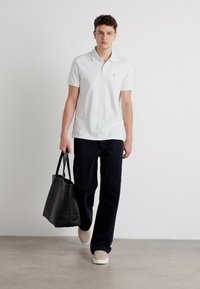 Hvid polo-shirt, mørke lige ben bukser, beige sneakers, bærende på en sort tote bag. Glat stof, afslappet pasform, minimalistisk design.
