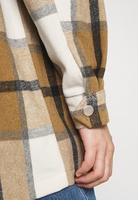 Camicia a quadri in tonalità di marrone, crema e grigio; presenta una texture morbida, polsini con bottoni e una vestibilità comoda con un orlo curvo.