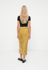 Donna con lunghi capelli biondi, vestita con una maglietta nera a maniche corte, una gonna midi dorata tempestata di paillettes e ballerine nere, in piedi di spalle.