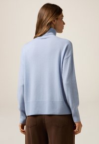 Maglione a collo alto di un blu chiaro in morbido tessuto testurizzato. Presenta una vestibilità comoda e orli e polsi a coste. Indossato con pantaloni marroni.