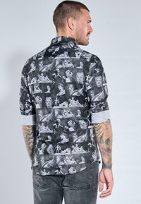 Schwarzes Hemd mit Knopfleiste, das eine Collage klassischer Statuen in Monochrom zeigt. Lange Ärmel mit aufgerollten Manschetten, entspannte Passform und abgerundeter Saum.