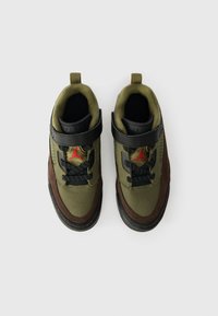 Ténis verdes e castanhos com uma faixa preta, sola de borracha e design de atacadores. Apresenta um logo vermelho e tecido texturizado para aderência.