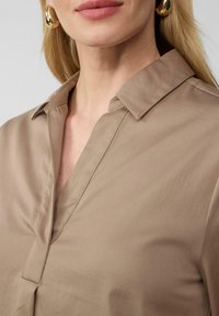 Blusa color beige con colletto e scollatura a V, texture liscia e dettaglio a tasca sul davanti. Sono visibili orecchini tondi dorati.