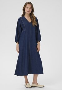 Ženska stoji oblečena v temno modro dolgo jeans obleko s tremi četrtinami rokavov in bež loaferji pred enostavno svetlo ozadje.